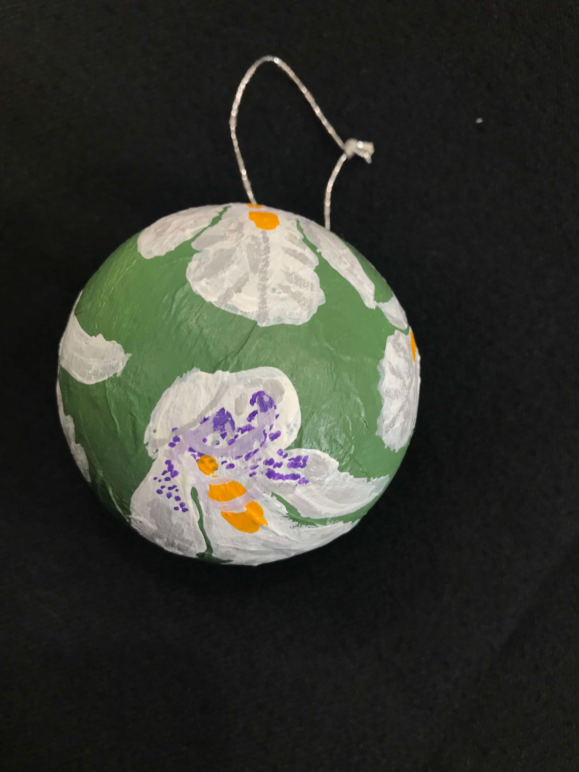 A round, hand-painted papier-mâché Christmas bauble with a floral pattern, predominantly green with white and purple flowers, and a red ribbon at the top.