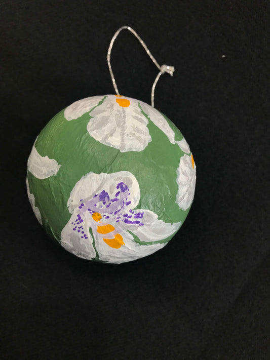 A round, hand-painted papier-mâché Christmas bauble with a floral pattern, predominantly green with white and purple flowers, and a red ribbon at the top.
